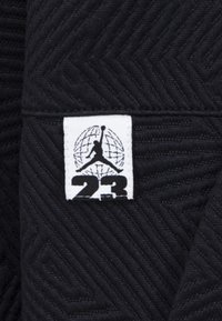 Černá texturovaná tkanina s vzorem herringbone. Bílá visačka zobrazuje logo se siluetou a pod ní je číslo "23".