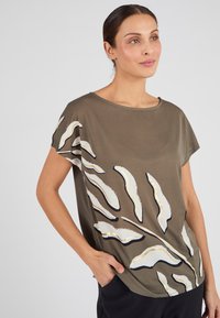 Mujer con una blusa verde oliva con patrones abstractos de hojas en blanco y negro, sonriendo mientras está de pie contra un fondo blanco liso.