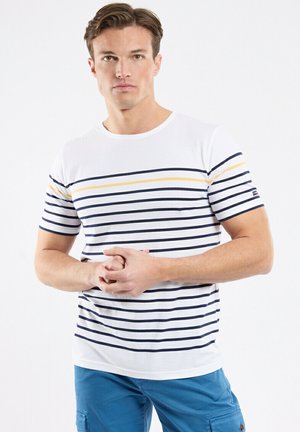 Man with short brown hair wearing white T-shirt with navy and yellow stripes and blue cargo shorts, standing with hands clasped.