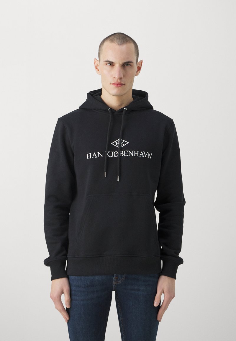 Črna kapuca s kenguru žepom, kapuco s vrvico, ter logotipom "HAN KØBENHAVN" v beli barvi. Ima rebraste manšete in okrasni rob.