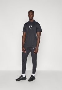 Czarna koszulka sportowa z krótkimi rękawami i logo, w zestawieniu z czarnymi spiczastymi spodniami i czarnymi butami sportowymi. Widoczne są białe skarpetki.