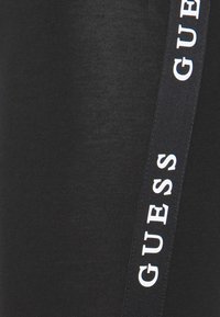 Czarne legginsy wykonane z gładkiego materiału, z bocznym paskiem z białym logo "GUESS". Projekt jest dopasowany i prosty.