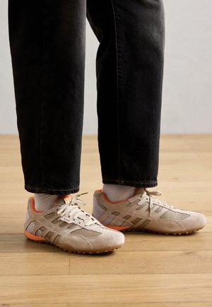 Beiga träningsskor i mesh och mocka, med orange detaljer och en texturerad gummisula. Bärs tillsammans med svarta byxor och vita strumpor.