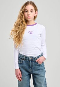 T-shirt bianco a maniche lunghe con scollo viola e testo grafico, abbinato a jeans in denim blu a vita alta con chiusura a bottone.