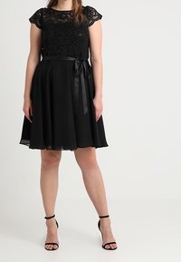 Donna che indossa un vestito nero a ginocchio con parte superiore in pizzo, nastro in vita e tacchi alti neri con cinturino alla caviglia, in piedi su uno sfondo bianco.