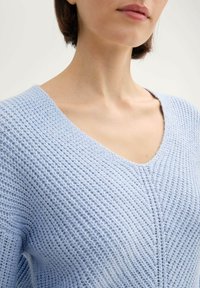 Lichtblauwe gebreide trui met een V-hals, textuurstof met een geribbeld patroon en zichtbare stiksels langs het midden.