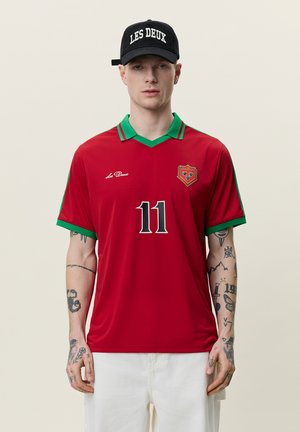 Mand iført rød og grøn sportstrøje nummer 11 og sort kasket med teksten "LES DEUX", viser tatoveringer på begge arme mod en ensfarvet baggrund.