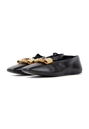 Zapatillas planas negras con punta y grandes lazos texturizados en color crema en la parte delantera, dispuestas una al lado de la otra sobre un fondo blanco.