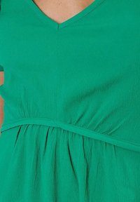 Groene jurk met een V-hals, gathered taille en zwierende stof met een gekreukt textuur. Het ontwerp heeft korte mouwen.