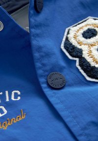 Chemise bleue avec un écusson de crâne brodé en fil blanc, noir et or. Comprend un bouton rond noir et des coutures contrastantes.