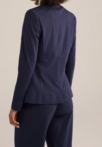 Woman wearing a fitted navy blazer and matching trousers, shown from the back against a plain neutral background.
