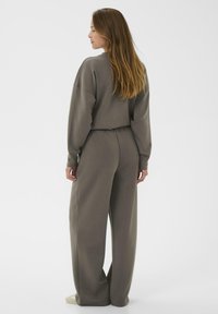 Woman with long hair stands facing away wearing loose, grayish-brown loungewear set with wide pants and matching sweatshirt on white background.