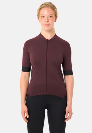 Jeune femme portant un maillot de cyclisme à manches courtes couleur bordeaux avec une fermeture éclair à l'avant et un cuissard noir, sur un fond uni.