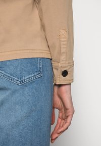 Béžová bavlněná bunda s knoflíkovou detailní na rukávu, spárovaná se světle modrými džíny, které mají diamantově tvarovanou zadní kapsu.