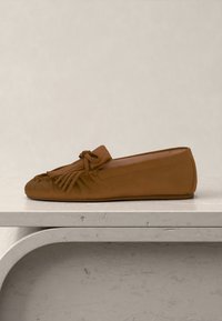 Mocassin en cuir marron avec des franges et un détail nœud sur le dessus, exposé sur une surface texturée de couleur claire.
