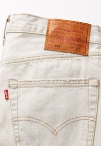 Poche arrière d'un jean Levi's 501 beige clair avec écusson de marque en cuir marron et étiquette en tissu rouge Levi's.