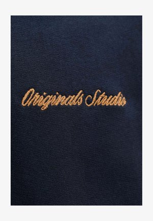 Mørk marineblåt stoff med en teksturert vev, med gullbrodert tekst som sier "Originals Studio" nær midten.