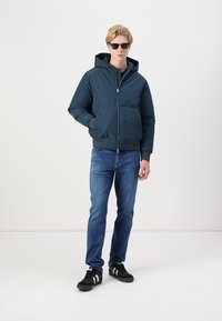 Chaqueta acolchada de color azul marino con capucha, cierre de cremallera y bolsillos laterales, combinada con vaqueros azules y zapatillas negras con detalles blancos.