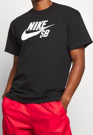 Osoba nosząca czarną koszulkę Nike SB z białym logo i jasnoczerwone szorty, ręce w kieszeniach, na jednolitym jasnym tle.