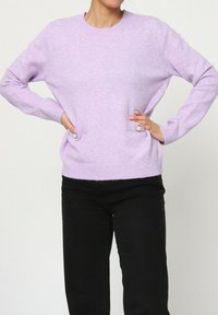 Pull en tricot lavande à manches longues, col rond, et deux poches latérales avec détails de boutons. Texture douce, coupe décontractée.