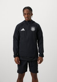 Mustat vedenpitävät takki, jossa on vetoketju, huppu ja valkoinen Adidas-logo. Rinnassa on Saksan maajoukkueen embleemi neljällä tähdellä.