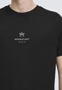 Černé bavlněné tričko s kulatým výstřihem a krátkými rukávy. Na hrudi je potištěné bílé logo a text: "KRONSTADT" a "CASUAL SINCE 1999."