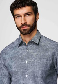 Homme barbu portant une chemise bleue à boutons avec un motif floral discret, regardant directement la caméra sur un fond uni.