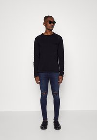 Suéter preto de mangas longas com decote redondo e bolso, emparelhado com jeans skinny rasgados azul-escuro e ténis pretos. Óculos de sol pretos usados.