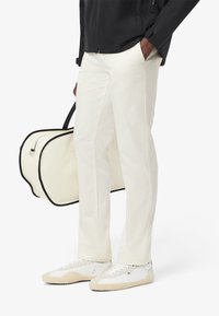 Pantaloni bianchi in cotone con un design a gamba dritta, abbinati a sneaker bianche e una borsa color crema con dettagli neri e una cerniera.