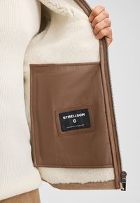 Interno di giacca in pelle marrone con fodera bianca in pile. Presenta una tasca in pelle marrone con un'etichetta nera con la scritta "STRELLSON" cucita sopra.