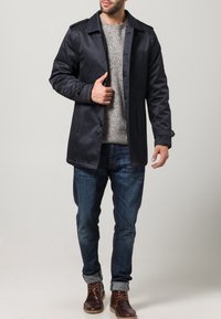 Marinblå trenchcoat med mjuk textur, knäppning med knappar och axelband, bärs över en ljusgrå stickad tröja och blå jeans.