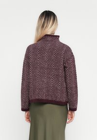 Woman with long hair wearing a burgundy chevron-patterned sweater and olive green skirt, viewed from behind against a plain background.