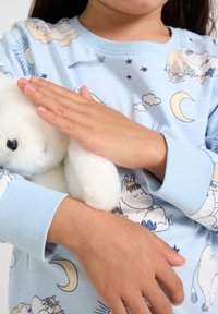 Lysebl�å pyjamasoverdel med tegneserieudskrifter af skyer, sol og måne. Manchetter ved ærmerne. Barnets hænder holder en fluffy hvid krammedukke.