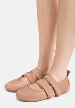 Füße in hellbraunen Wildleder- und Lederschuhen mit zwei breiten Klettverschlüssen, vor einem schlichten hellen Hintergrund dargestellt.