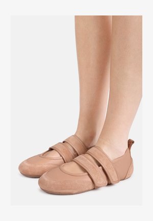 Voeten die lichtbruine schoenen van suède en leer dragen, met twee brede klittenbandsluitingen, afgebeeld tegen een effen lichte achtergrond.