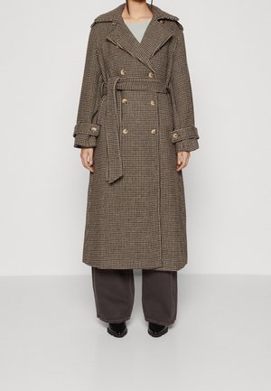 Lange dubbelgeknoopte trenchcoat in bruin en beige ruitpatroon, met riem, gedragen door persoon met wijde, donkere broek en zwarte schoenen.
