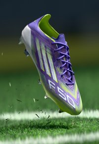 Purple and lime green soccer cleat with textured synthetic upper, featuring prominent white stripes and clear studs, shown hovering above grass.