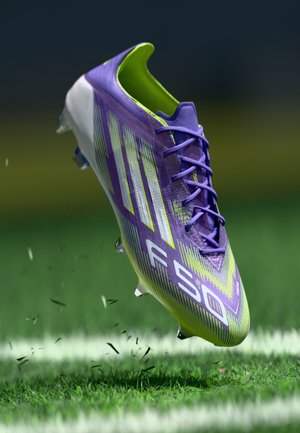 Purple and lime green soccer cleat with textured synthetic upper, featuring prominent white stripes and clear studs, shown hovering above grass.