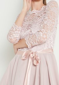 Woman wearing a pale pink lace dress with three-quarter sleeves and a satin ribbon tied in a bow at the waist.