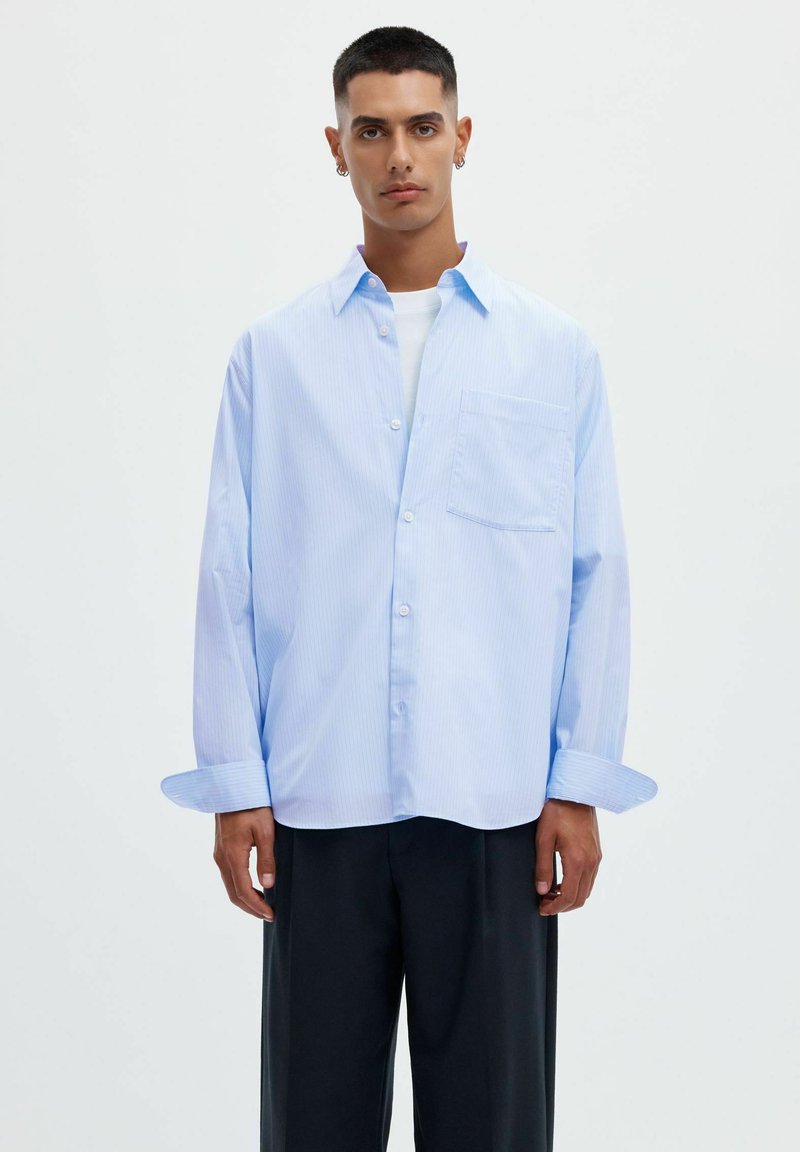 Jeune homme aux cheveux courts porte une chemise oversize bleu clair boutonnée par-dessus un t-shirt blanc et un pantalon noir à jambes larges sur un fond uni.