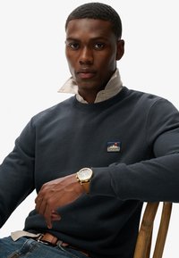 Marineblauwe fleece sweatshirt met een ronde hals en een logo patch. Het model draagt een beige overhemd eronder en een gouden horloge. Spijkerbroek zichtbaar.