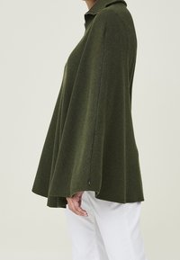 Poncho a maniche lunghe in maglia verde scuro, con colletto alto, silhouette fluida e dettagli di cuciture sottili.