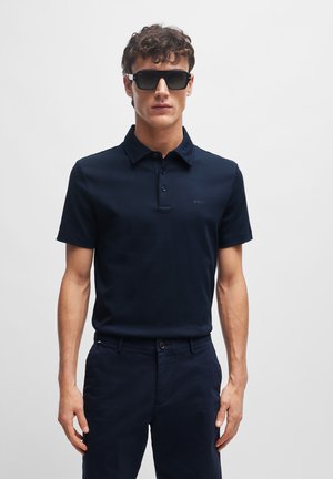 Marineblauwe polo met een drieknopensluiting, korte mouwen en een subtiele merklogo op de borst. Het model draagt een donkere zonnebril.