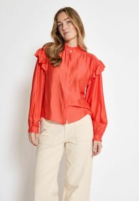 Blusa di seta rossa con colletto arricciato e maniche arricciate, chiusura con bottoni e polsini elasticizzati. Abbinata a pantaloni ampi color crema.