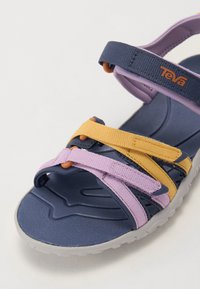 Teva sandales tumši zila, lavandas un dzeltenā krāsā. Iespējas: regulējamas austošanās siksnas, strukturēta zole un pelēka gumijas zole.