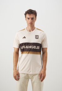 Maglietta da calcio beige con una striscia orizzontale marrone, caratterizzata da dettagli dorati, con il testo "LOS ANGELES" e un logo sul lato sinistro del petto.