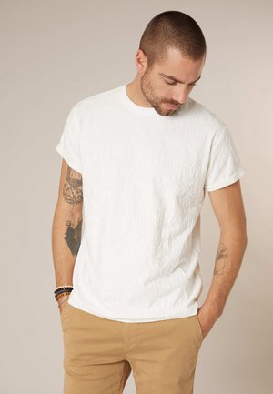 Homme aux cheveux courts et tatouages portant un t-shirt blanc texturé et un pantalon beige, regardant vers le bas les mains dans les poches.