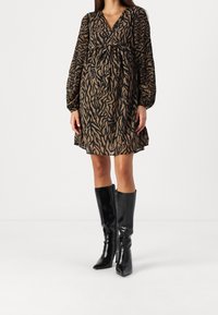 Žena v hnědočerných šatech s potiskem zvířecí srsti, délky po kolena, s dlouhými průhlednými rukávy a černými vysokými kozačkami na bílem pozadí.