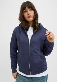 Felpa con zip di colore navy, realizzata in materiale morbido, con cappuccio, tasche frontali e un piccolo dettaglio con logo. Indossata sopra una t-shirt bianca.