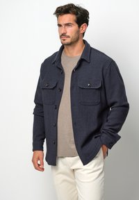 Marineblaues gewobenes Hemdjackett mit zwei Brusttaschen, Knopfverschluss und strukturierter Textur. Getragen über einem beigen Rundhalshemd.